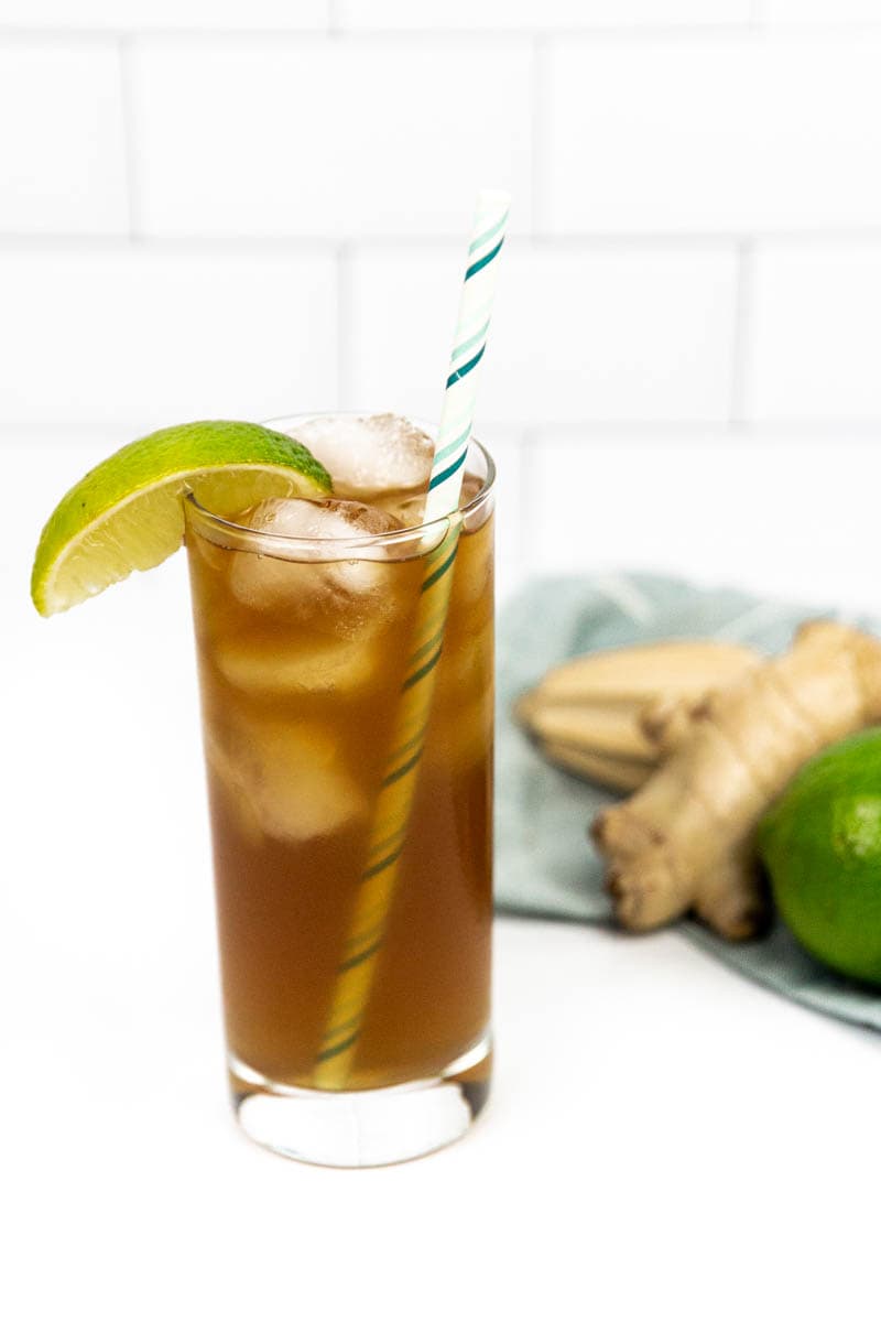A tall glass holds a dark tan cocktail with ice, garnished with a green striped straw and lime wedge. Ginger and a lime sit in the background on a light blue dishtowel