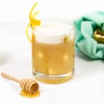 A honey lemon whiskey sour cocktail sits with several of its ingredients with a cocktail jigger on a teal dish towel in background