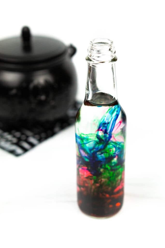 blue, green and red food coloring swirling together in closeup shot of glass bottle of vodka with out of focus cauldron in background