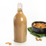 almond syrup in glass bottle with black bowl of almonds on green dish towel and several scattered almonds