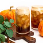 two glasses of iced pumpkin cream cold brew with decorative pumpkins, green leaves, cinnamon sticks and a gold and black spoon on white background