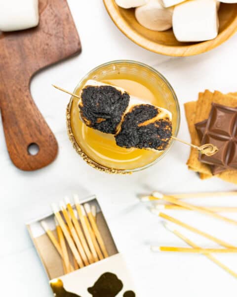 overhead view of s'mores old-fashioned cocktail with matches, a stack of graham crackers and chocolate and marshmallows scattered around