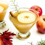 Two apple cider margarita cocktails next to two red apples, some fall leaves and rosemary sprigs on a white background.