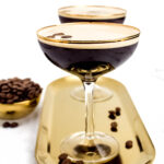 A shiny gold tray sits on a white marble surface holding two martini glasses filled with coffee-colored liquid. Coffee beans are scattered around