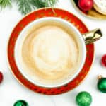 A festive cookie latte sits on a red and gold plate with green and red holiday ornaments surrounding it.