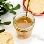 A jar of sugar cookie simple syrup sits on a white marble surface with a gold spoon in the jar. There are cookies dusted with sugar set around it and holiday foliage.