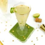 A champagne version of a moscow mule in a glass rests on a green coaster surrounded by gold cocktail tools and half a lime.
