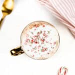 Overhead view of a foamy coffee beverage garnished with crushed candy canes. Additional candy canes and a gold spoon sit alongside.