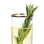 Rosemary sprigs stick out of a gold-rimmed glass with a bubbly cocktail inside.