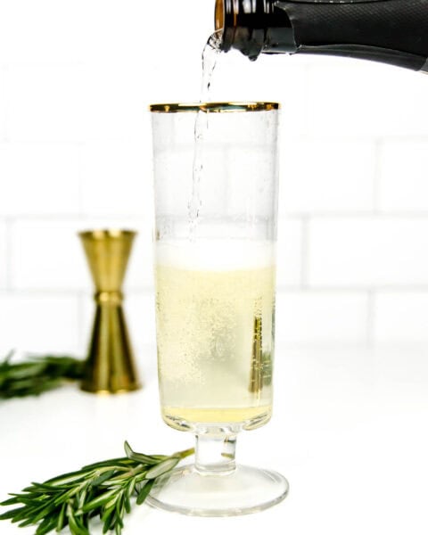 Champagne is poured into a short-stemmed flute with rosemary sprigs and a gold cocktail jigger alongside on a white background.