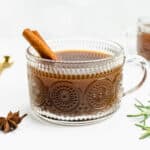 A glass mug holds a warm cider drink surrounded by whole spices and rosemary sprigs.