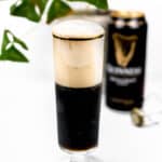 A glass is half full of dark liquid while foam fills the remaining glass. A can of Guinness sits out of focus in the background along with the cage from a champagne bottle.