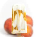 An orange and white swirled popsicle is held in front of orange peaches.