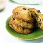 A stack of three cookies with chocolate chips sit on a green plate, while another cookie with a bite missing leans on the stack.