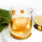 An old-fashioned cocktail made with Irish whiskey sits alongside a gold cocktail jigger resting on a green dish towel and a stack of gold coasters.