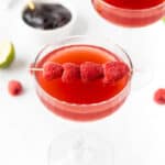 A row of raspberries on a toothpick garnish a light red cocktail.