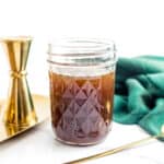 A jar of Irish Cream syrup sits next to a gold plate holding a gold bowl of cocoa powder and a gold cocktail jigger. A gold spoon sits on a green dish towel to the side.