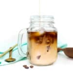 Irish cream is poured into a glass mug of iced coffee. Behind the mug sits a teal dish cloth with a gold straw and spoon resting on top and a brown scoop of coffee beans.