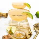 Two champagne margaritas sit on a gold tray along with lime wedges and a champagne cork.