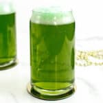 A can-shaped glass of green beer sits in front of another glass and gold bead necklaces.