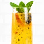 A closeup view of a glass with gold stars containing a sparkling yellow beverages. A peach slice, blackberry and mint sprig garnish the glass.
