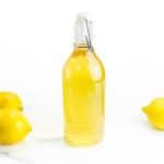 A bottle of yellow liqueur sits next to three whole lemons on a white marble surface.