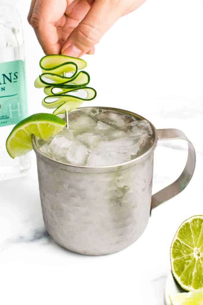 a hand adds a cucumber ribbon garnish on a cocktail pick to a silver mug.