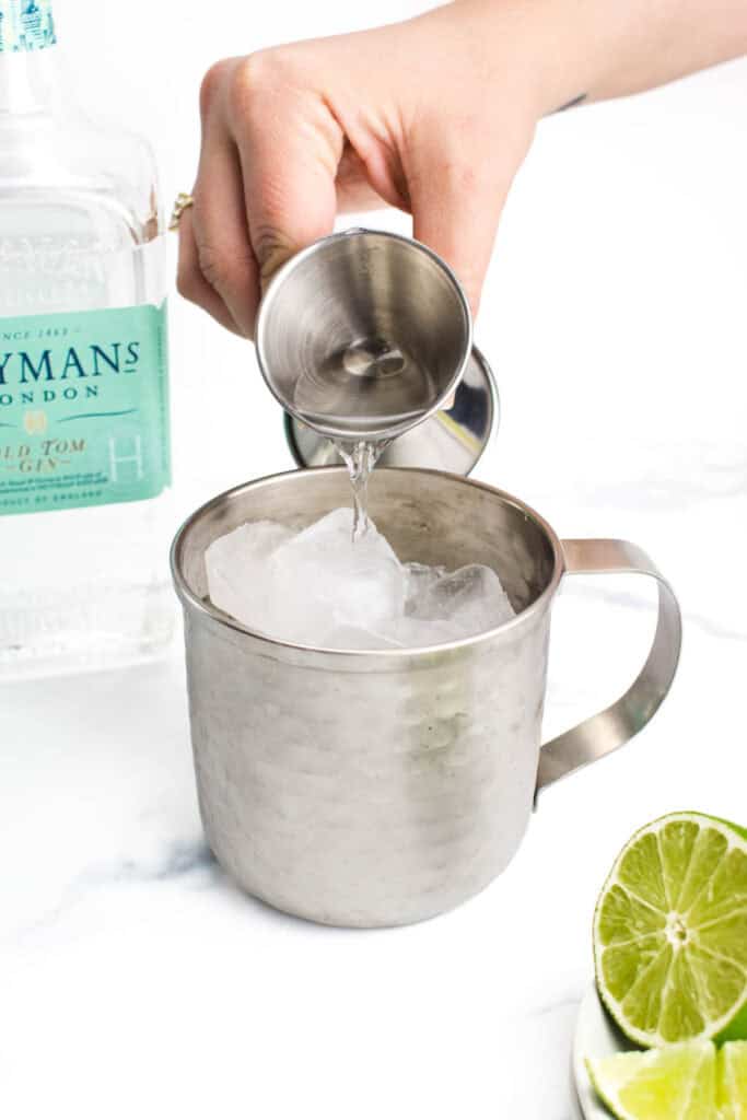 a hand holding a jigger pours old tom gin into a mule mug.