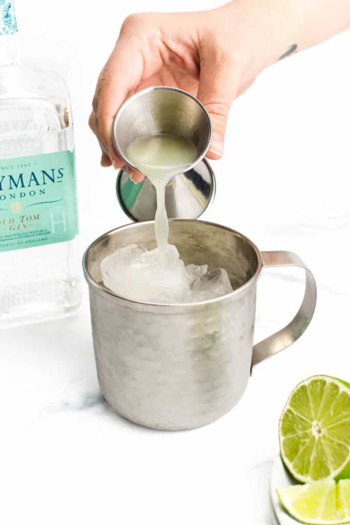 a hand pours lime juice from a jigger into a silver mug filled with ice.