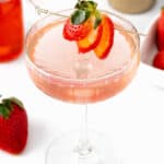 a sliced strawberry on a gold cocktail pick over a pink cocktail.