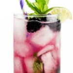 A Blackberry Mojito is garnished with a sprig of mint, lime wedge, whole blackberry and striped purple and white straw.