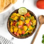 A wooden spoon sits on a white marble surface next to a bowl of summery salsa. Other ingredients and chips sit on the same surface.