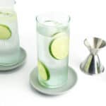limes, a silver jigger and two vodka sodas on grey coasters.