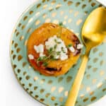 A baked peach half with honey and goat cheese on a gold speckled plate with a gold spoon on a white background.