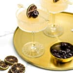 A gold tray holds two rum cocktails in coupe glasses, the lid to a cocktail shaker and a bowl of cherries. additional garnishes and tools sit beside on a white marble surface.