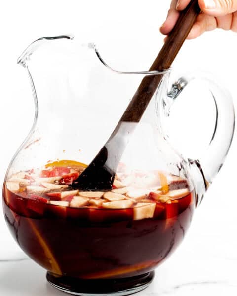 A wooden spoon stirs fruit and red wine sangria in a clear glass pitcher