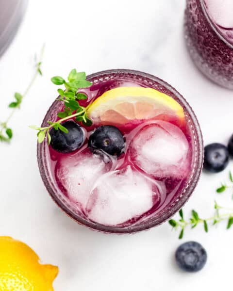 An overhead view of a lemon wedge, blueberries and herb sprig alongside ice in a purple drink.