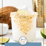 A glass of white coconut margarita with toasted coconut on the rim, garnished with a lime wedge, surrounded by fresh coconut, lime slices, and a cocktail shaker.