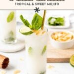 A glass of coconut mojito garnished with lime, mint, and coconut flakes, with a gold straw, sits on a white surface next to fresh ingredients and another vibrant coconut mojito.