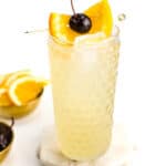 A John Collins cocktail sits next to gold bowls of cherries and orange slices.