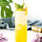 A tall glass of yellow mango mojito garnished with mint, a lime slice, and a straw. Two purple flowers and a piece of lime are placed nearby on the white surface, with green leaves and a blue cloth in the background.