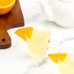Two tea shots in clear mugs garnished with orange wedges next to a white teapot.