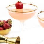 A Strawberry Daiquiri garnished with a strawberry on a skewer sits next to a bowl of strawberries and a gold cocktail jigger.