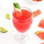 A refreshing watermelon drink sits in a glass surrounded by lime slices, watermelon slices and watermelon chunks.