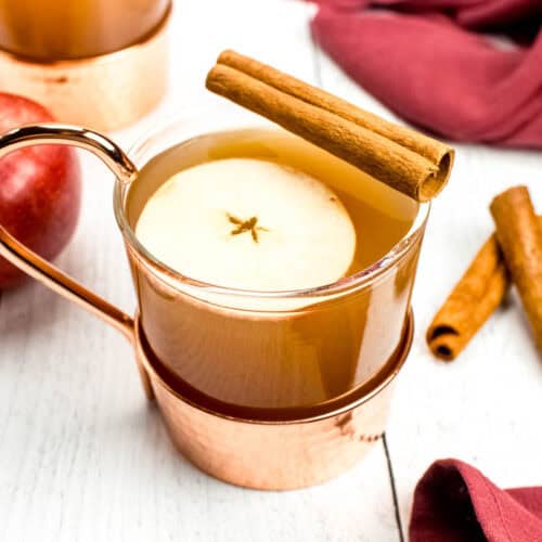 A cup of apple cider with cinnamon sticks and apples.