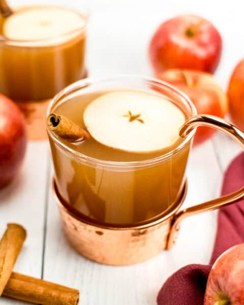 A cup of apple cider with cinnamon sticks and apples.
