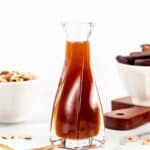 A glass bottle of toffee sauce next to a bowl of nuts and a spoon.