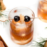 A Christmas Old-Fashioned cocktail in a glass decorated with star patterns, garnished with two cherries and a slice of fruit on a skewer, placed on a white surface with greenery and cookies nearby.