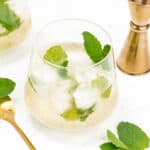 Two glasses of iced Hugo Spritz with gold-colored utensils on a white surface.