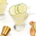 Two cucumber garnished cocktails with a golden cocktail shaker on a marble countertop.